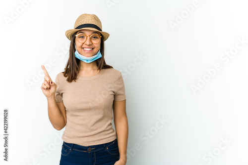 Young latin woman wearing hat and mask to protect from covid isolated on white background showing number one with finger.