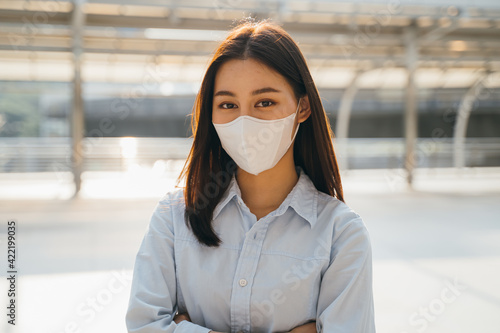 Portrait of young Asian businesswoman wearing covid-19 protective face mask for safety against coronavirus with crossed arms outside office