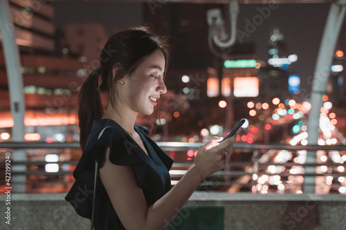 Side-view of beautiful young Asian happy woman standing over footbridge checking smartphone and browsing through apps while checking email