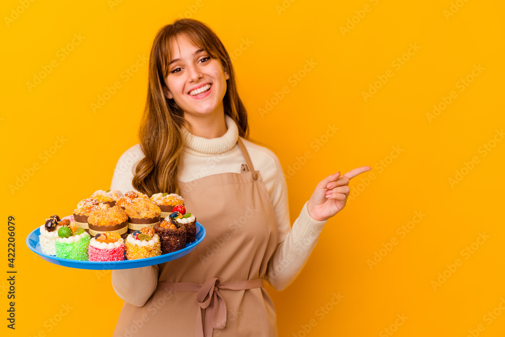 Young pastry chef woman isolated on yellow background smiling and pointing aside, showing something at blank space.