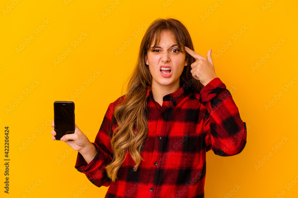 Young caucasian woman holding phone isolated on yellow background showing a disappointment gesture with forefinger. Young caucasian woman holding phone isolated on yellow background showing a disappointment gesture with forefinger.
