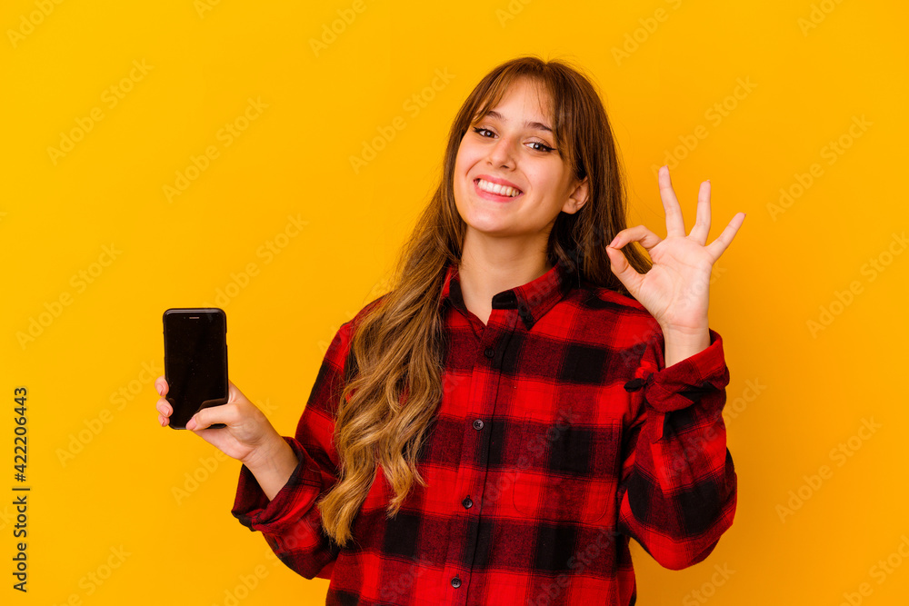 Young caucasian woman holding phone isolated on yellow background cheerful and confident showing ok gesture. Young caucasian woman holding phone isolated on yellow background cheerful and confident showing ok gesture.