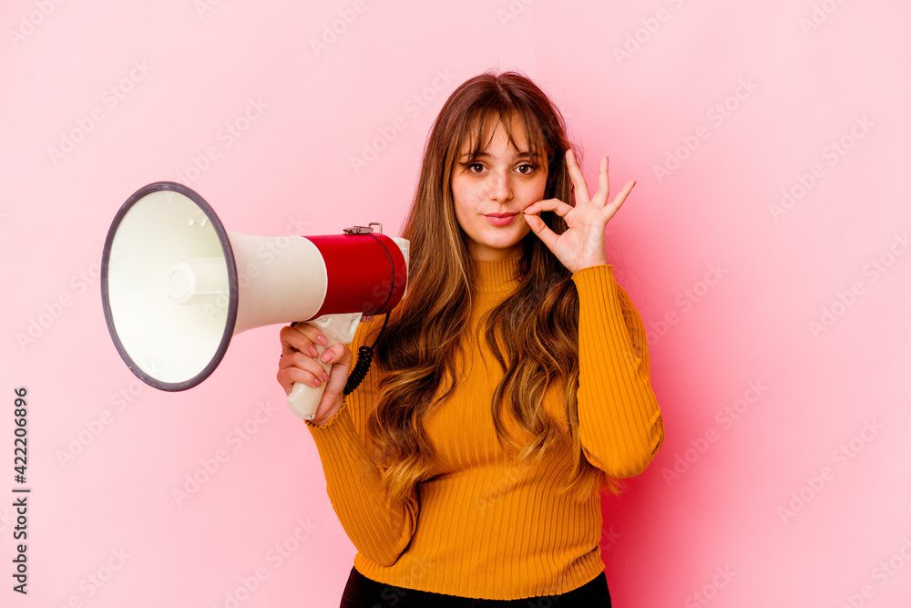Young caucasian woman holding a megaphone isolated with fingers on lips keeping a secret. Young caucasian woman holding a megaphone isolated with fingers on lips keeping a secret.