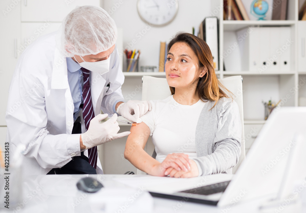 Doctor in white coat and protective mask giving vaccine injection to woman manager in office