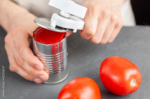 A woman is carefully opening a can of tomato paste on a kitchen counter using a white plastic can opener. She is preparing a meal for which she uses both fresh and pureed preserved tomatoes photo