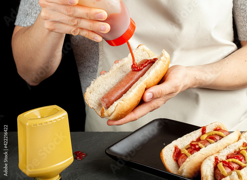 A caucasian woman is preparing hot dog sandwiches. She is squeezing tomato ketchup from plastic bottle  on to a beef sausage inside a hot dog bun. Other sandwiches are on plate over the counter. photo