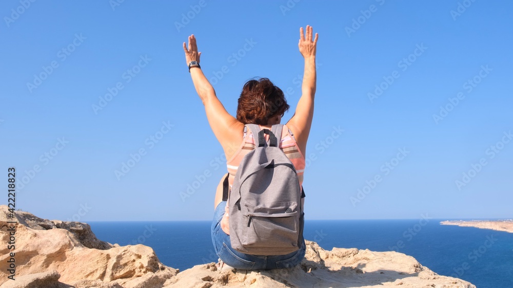 young happy woman with a backpack sits on a rock with her hands raised and looks at the valley below. The concept of freedom and ease. young happy woman with a backpack sits on a rock with her hands raised and looks at the valley below. The concept of freedom and ease.