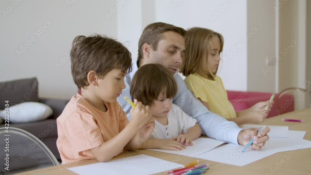 Focused dad drawing with marker on paper and lovely kids sitting near him. Caucasian father painting with pen and playing with children in living room. Side view. Childhood and family time concept Focused dad drawing with marker on paper and lovely kids sitting near him. Caucasian father painting with pen and playing with children in living room. Side view. Childhood and family time concept