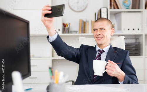 Young smiling cheerful businessman in white office making selfie on smartphone