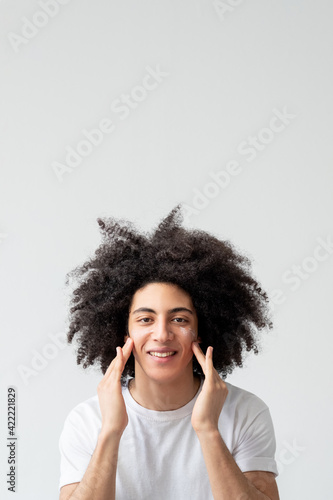 Man skincare. Cosmetic product. Skin spf protection. Happy cheerful handsome guy with long curly hair applying face cream smiling isolated on light empty space advertising background.
