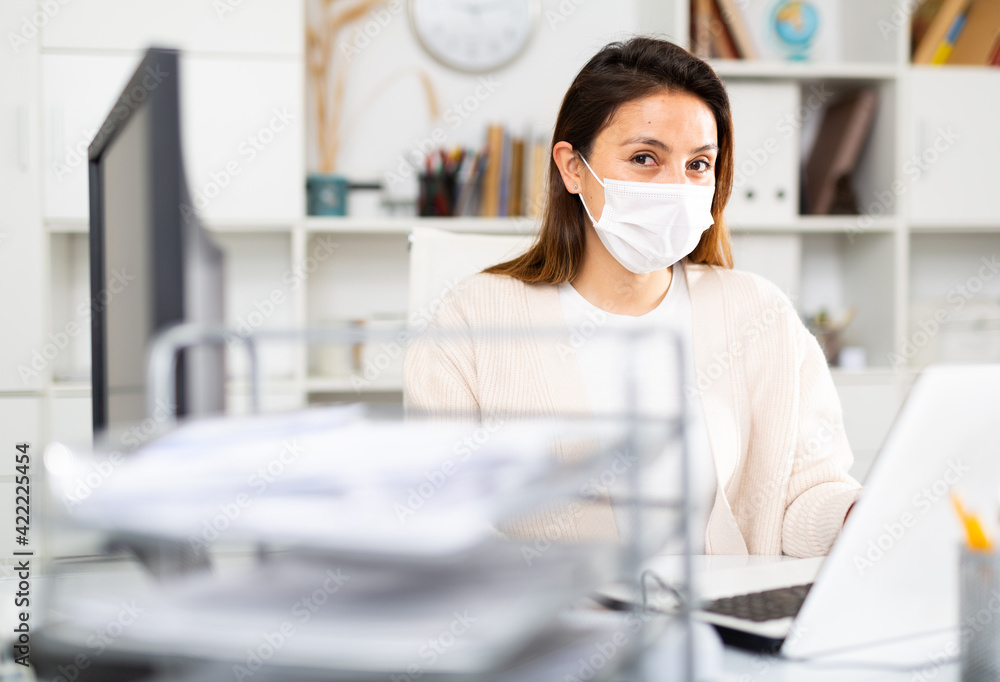 Secretary girl in a protective mask during a pandemic sits at her workplace in the office in front of a laptop