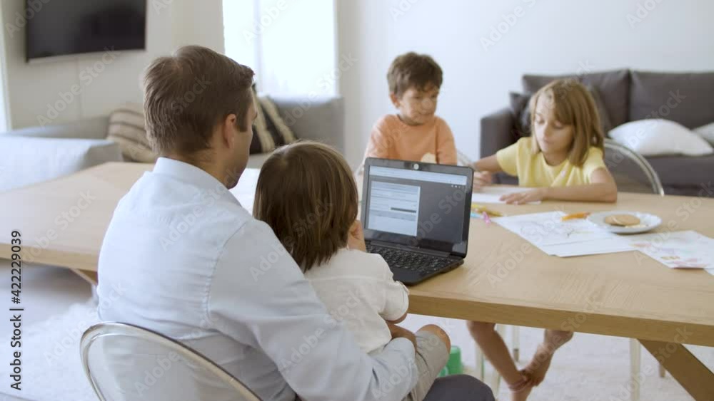 Freelance dad working from home, using laptop, holding child on lap, looking after kids playing in living room. Distance job, fatherhood concept