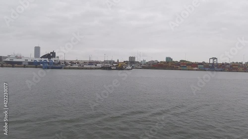 Ship sails along shore past lighthouse and port facilities with cranes yachts boats on rippling water under cloudy sky timelapse