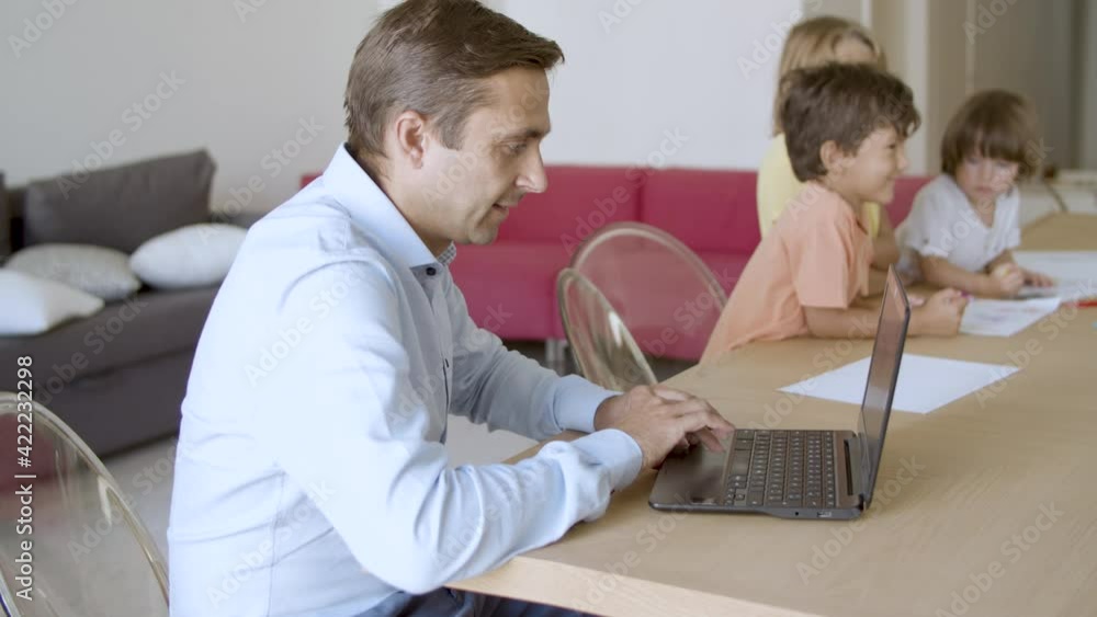 Caucasian dad working on laptop and cute kids painting doodles at table. Concentrated blonde girl drawing with marker and brother looking at her. Static camera. Childhood and weekend concept Caucasian dad working on laptop and cute kids painting doodles at table. Concentrated blonde girl drawing with marker and brother looking at her. Static camera. Childhood and weekend concept