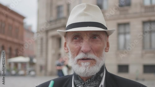 Senior man with grey beard and blue eyes in hat and satin handkerchief around neck talks against historical buildings closeup