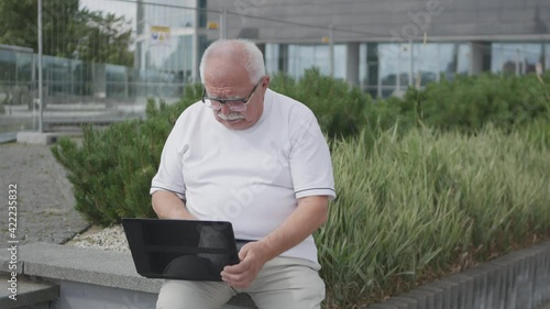 Senior man with glasses in white T-shirt sits on bench holding laptop and types message against green bushes and office closeup