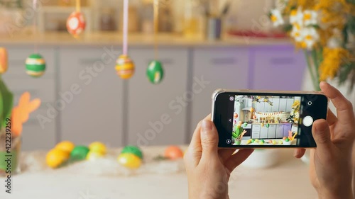 Hands hold smartphone. On display photo composition for holiday of spring Happy Easter. Girl takes pictures on phone painted eggs on Easter decorated on table of the house. 