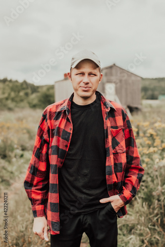 a man in a plaid red shirt and a cap. A man living in nature. Farmer on the background of a wooden house. American guy in the field
