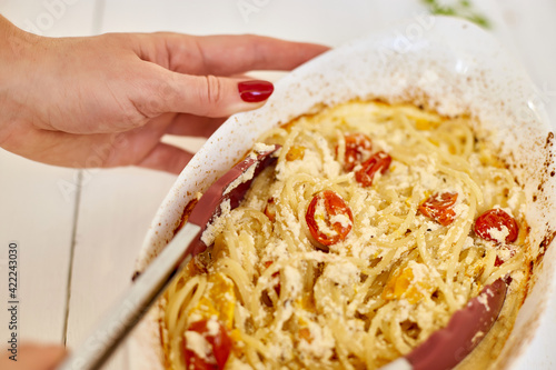 Woman hand's cooking trending viral baked tomatoes and feta with pasta, Fetapasta on a white wooden background . New, popular dinner dish, Top view, copy space.. photo