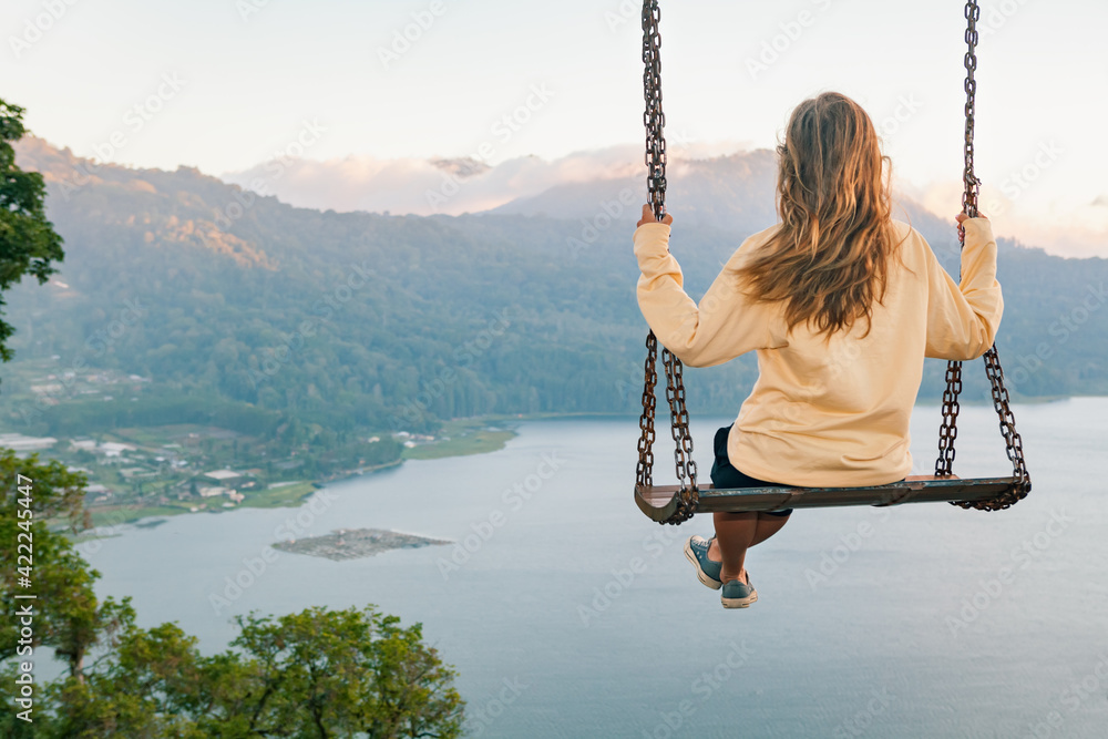 Summer vacation. Young woman sit on tree rope swing on high cliff above tropical lake. Happy girl looking at amazing jungle view. Buyan lake is popular travel destinations in Bali island, Indonesia