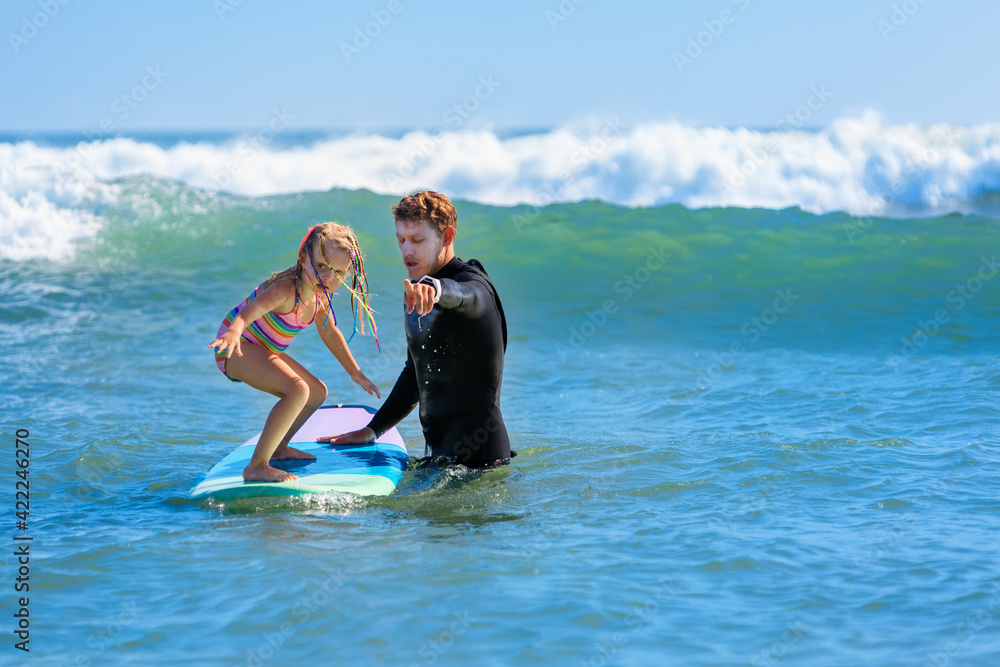 © Tropical studio - Little surf girl - young surfer learn to ride on surfboard with instructor at surfing school. Active family lifestyle, kids water sport lessons, swimming activity in summer camp. Vacation with child.