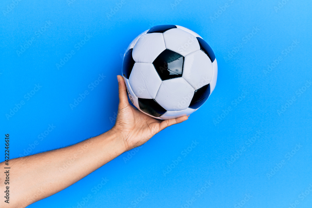 Hand of young hispanic man holding soccer ball over isolated blue background.