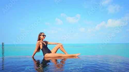  Young Woman in Black Swimming Suit Sitting on the Edge of Infinity Pool on Turquoise Seascape Background Wearing Sunglasses on a Sunny Day Slow Motion
 photo