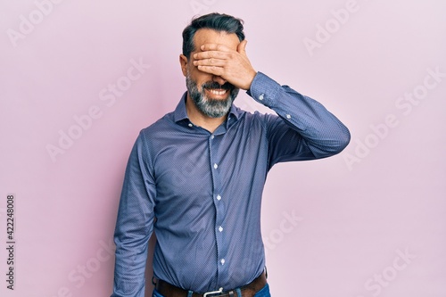 Middle age man with beard and grey hair wearing business clothes smiling and laughing with hand on face covering eyes for surprise. blind concept.