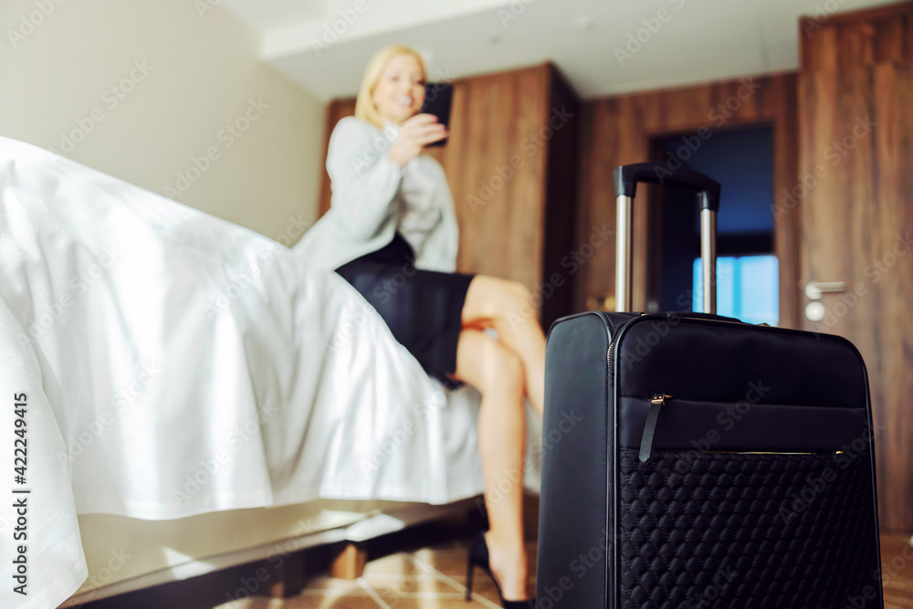 Closeup of suitcase in hotel room. In the background is a businesswoman sitting on the bed and using a smart phone. She is at the symposium.