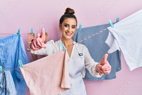 Beautiful brunette young woman washing clothes at clothesline approving doing positive gesture with hand, thumbs up smiling and happy for success. winner gesture.