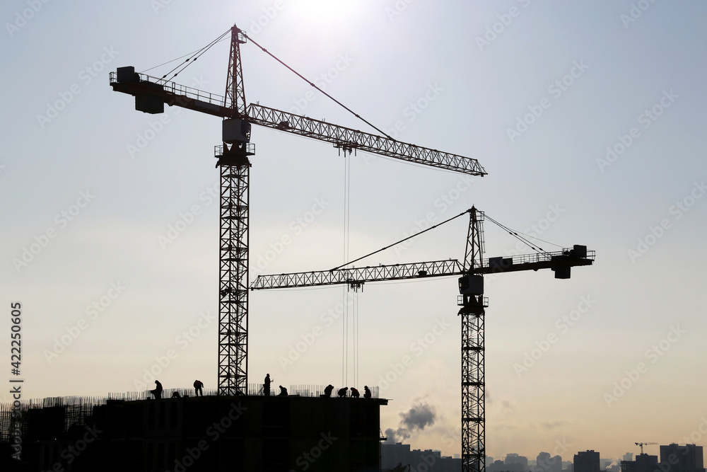 Silhouettes of construction cranes and workers on unfinished residential building against sunshine. Housing construction, apartment block in city