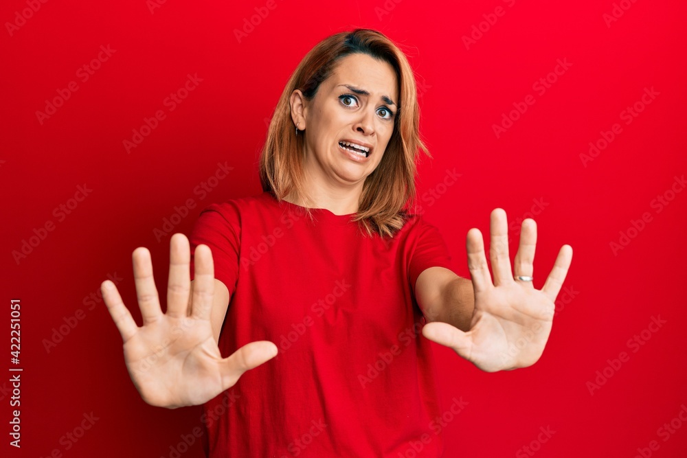 Hispanic young woman wearing casual red t shirt afraid and terrified with fear expression stop gesture with hands, shouting in shock. panic concept. Hispanic young woman wearing casual red t shirt afraid and terrified with fear expression stop gesture with hands, shouting in shock. panic concept.