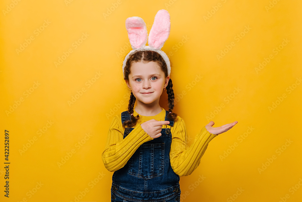 Horizontal shot of happy preschool girl in pink bunny fluffy ears, points for finger aside, demonstrates something over blank space, isolated over yellow studio background. Easter holiday concept