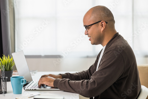 Indian man using laptop sitting in living room texting on laptop sending message or chatting with online social media.