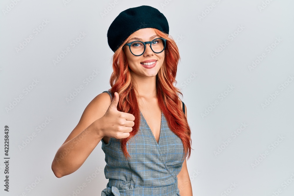 Young redhead woman wearing fashion french look with beret doing happy thumbs up gesture with hand. approving expression looking at the camera showing success. Young redhead woman wearing fashion french look with beret doing happy thumbs up gesture with hand. approving expression looking at the camera showing success.