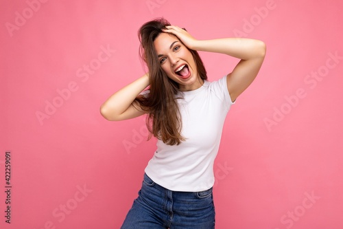 Young beautiful brunet woman. Trendy lady in casual summer white t-shirt for mock up. Positive female shows facial sincere emotions. Funny model isolated on pink background with free space