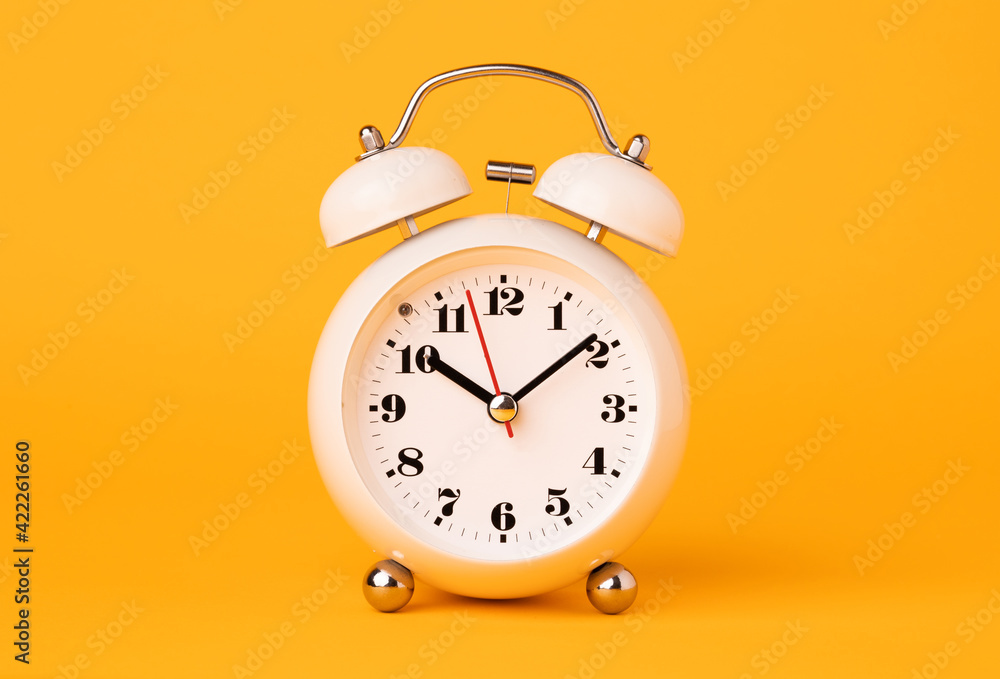 Small white alarm clock, black numbers, set the time placed on a table. Clock on isolated yellow background. Small white alarm clock, black numbers, set the time placed on a table. Clock on isolated yellow background.