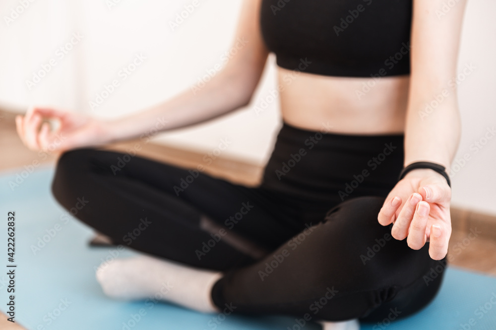 Close-up of the hands of a girl sitting on a yoga mat in a tracksuit and meditating in the lotus position. Faceless, young woman. Concentration, relaxation, mental health at home.
