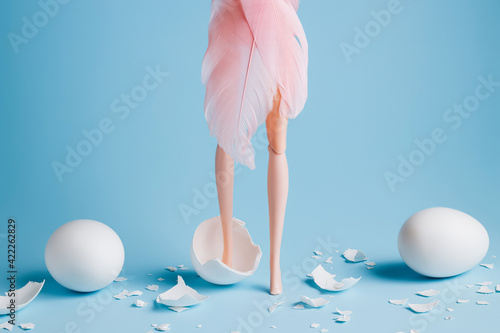 Girl doll with elegant pink feather dress legs among cracked white eggs on pastel blue background. Creative minimal Easter or fashion concept. photo