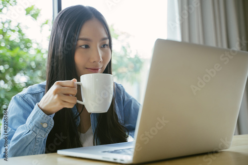 Asian woman using a laptop working at home hold a coffee cup while sitting at work.