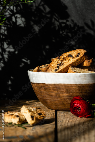 Homemade almond and chocolate biscotti with glass of coffee and ranunculus flowers on wooden table on dark background.