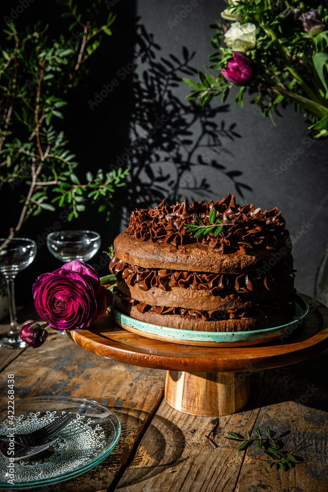 Homemade chocolate and cherry cake on wooden stand with flowers on dark background. Homemade chocolate and cherry cake on wooden stand with flowers on dark background.