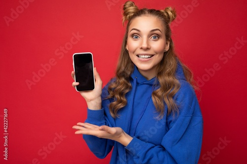 Attractive smiling young blonde woman wearing stylish blue hoodie isolated on red background with copy space holding smartphone showing phone in hand with empty screen display pointing at gadjet