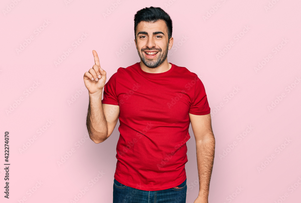 Hispanic man with beard wearing casual red t shirt showing and pointing up with finger number one while smiling confident and happy. Hispanic man with beard wearing casual red t shirt showing and pointing up with finger number one while smiling confident and happy.