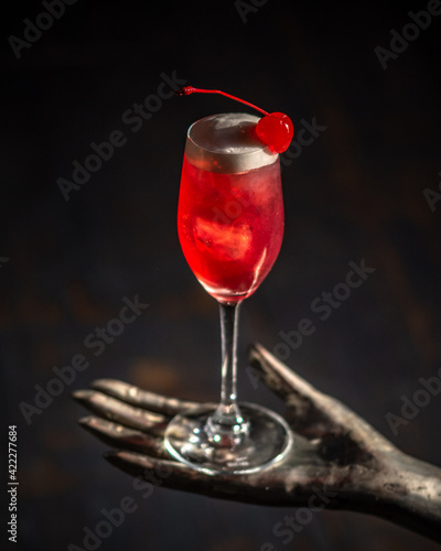bicchiere da cocktail lungo con liquore rosso, schiuma d'uovo e ciliegia rossa sciroppata al maraschino poggiato su scultura a forma di mano di ferro