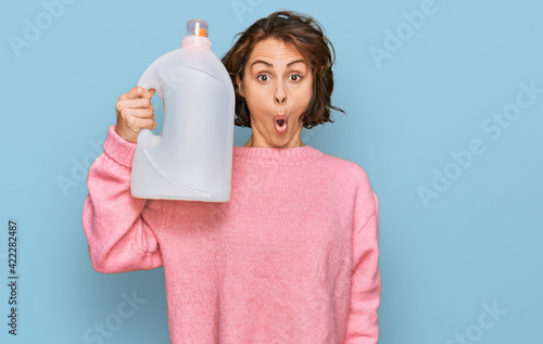 Young hispanic woman doing laundry holding detergent bottle scared and amazed with open mouth for surprise, disbelief face