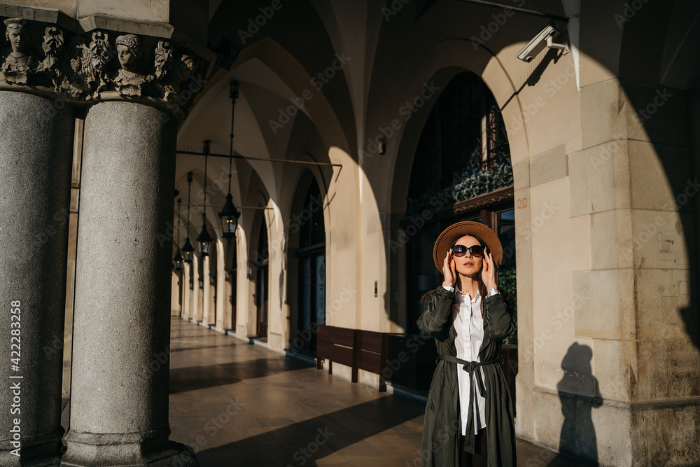 Outdoor fashion portrait of elegant, luxury woman wearing beige hat, sunglasses, trendy white shirt, in a green trench coat, walking in street of European city. Copy, empty space for text