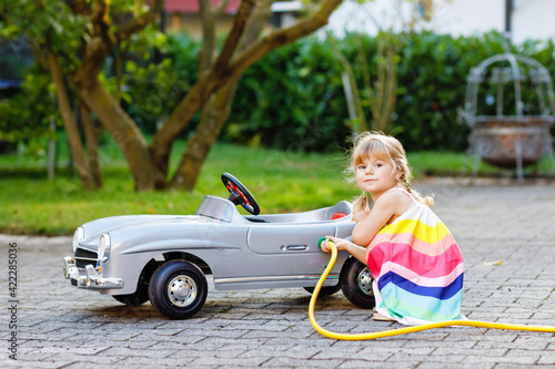 Little toddler girl playing with big vintage toy car and having fun outdoors in summer. Cute child refuel car with water. Girl using garden hose and fill up with gasoline, role game gas station.