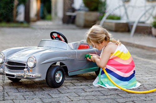 Little toddler girl playing with big vintage toy car and having fun outdoors in summer. Cute child refuel car with water. Girl using garden hose and fill up with gasoline, role game gas station.