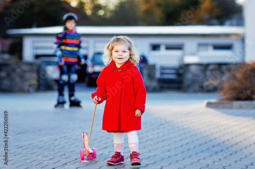 Outdoor portrait of little cute toddler girl in red coat aon spring sunny day with push wooden toy. Healthy happy baby child walking in the city. Brother boy on roller skating on background photo